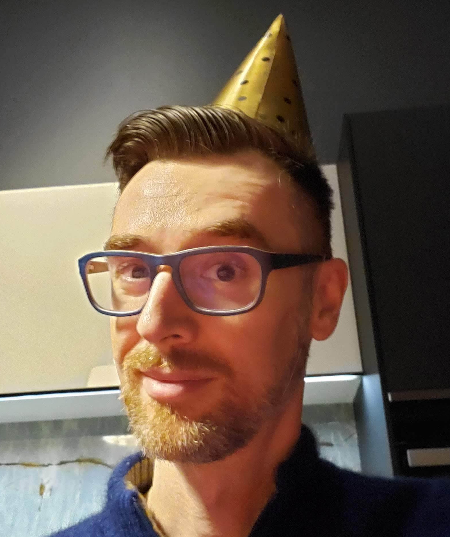A white man in blue glasses wearing a New Year's hat.